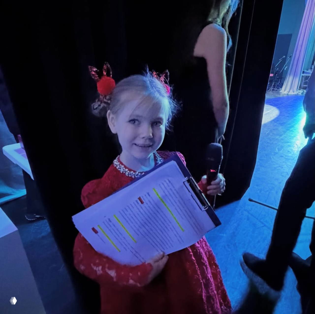 Девочка в красном платье за кулисами держит папку с подсвеченным текстом и микрофон, готовится выйти на сцену школьного праздничного концерта