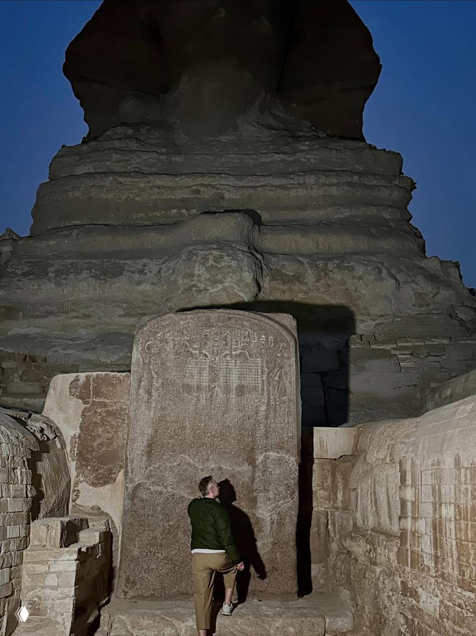 Ночной кадр: человек стоит у древней стелы под силуэтом Сфинкса, искусственное освещение подчёркивает рельефы и надписи на камне.