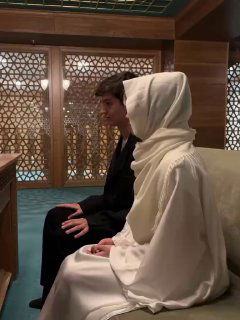 Крупный план пары в мечети: молодая женщина в белом одеянии и мужчина сидят рядом на ковре, спокойная интимная сцена религиозного обряда.