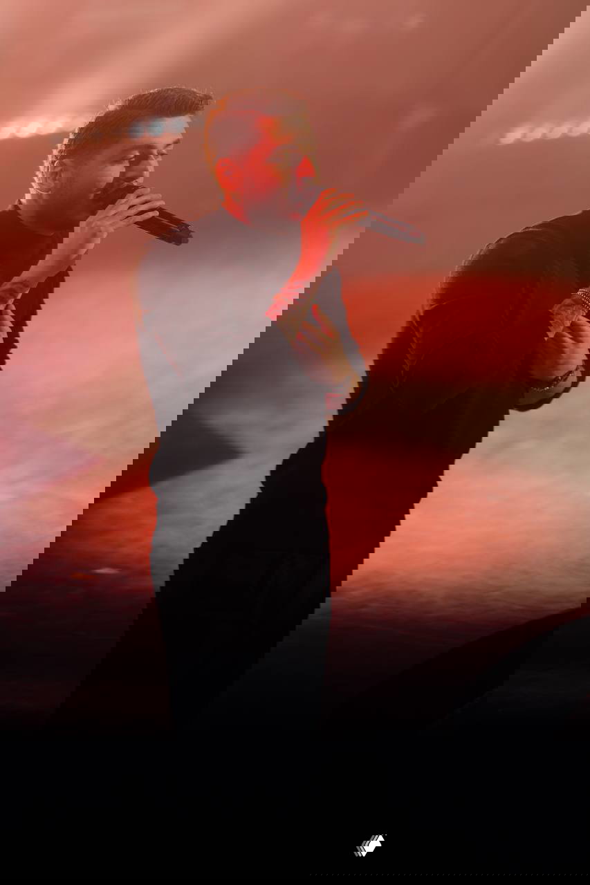 Концертная фотография: турецкий певец BLOK3 поёт в микрофон на сцене при тёплой красной подсветке, артист эмоционально держится за грудь.