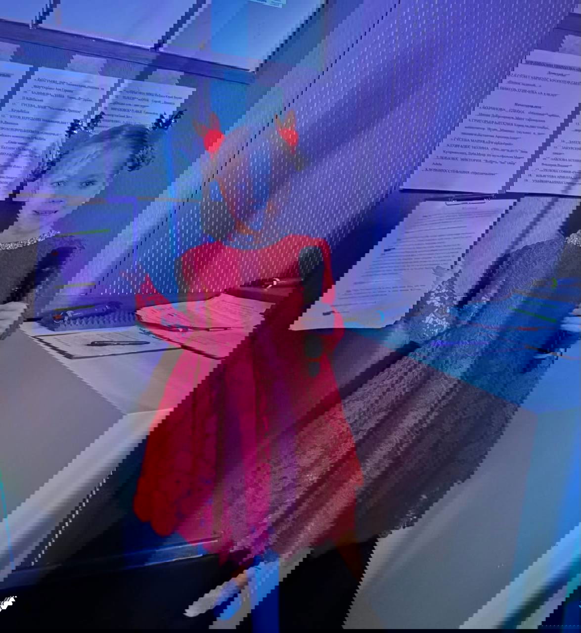 Девочка в красном кружевном платье улыбается у кулис, держит микрофон и распечатки со сценарием; подготовка к школьному концерту и репетиция