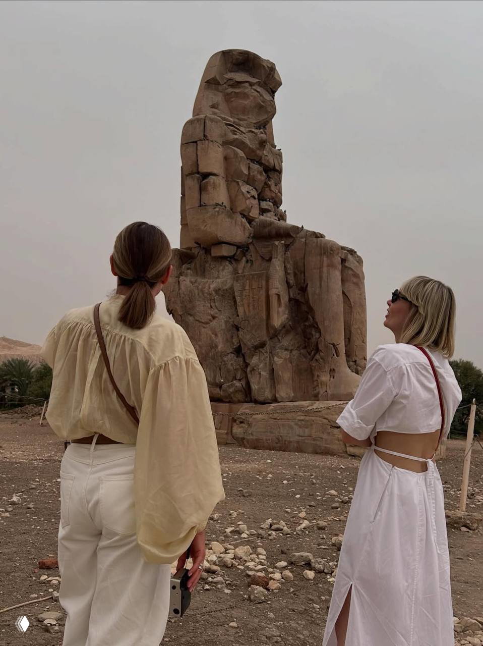 Две женщины в лёгкой одежде рассматривают массивную древнюю статую на фоне песчаного ландшафта и редкой растительности.