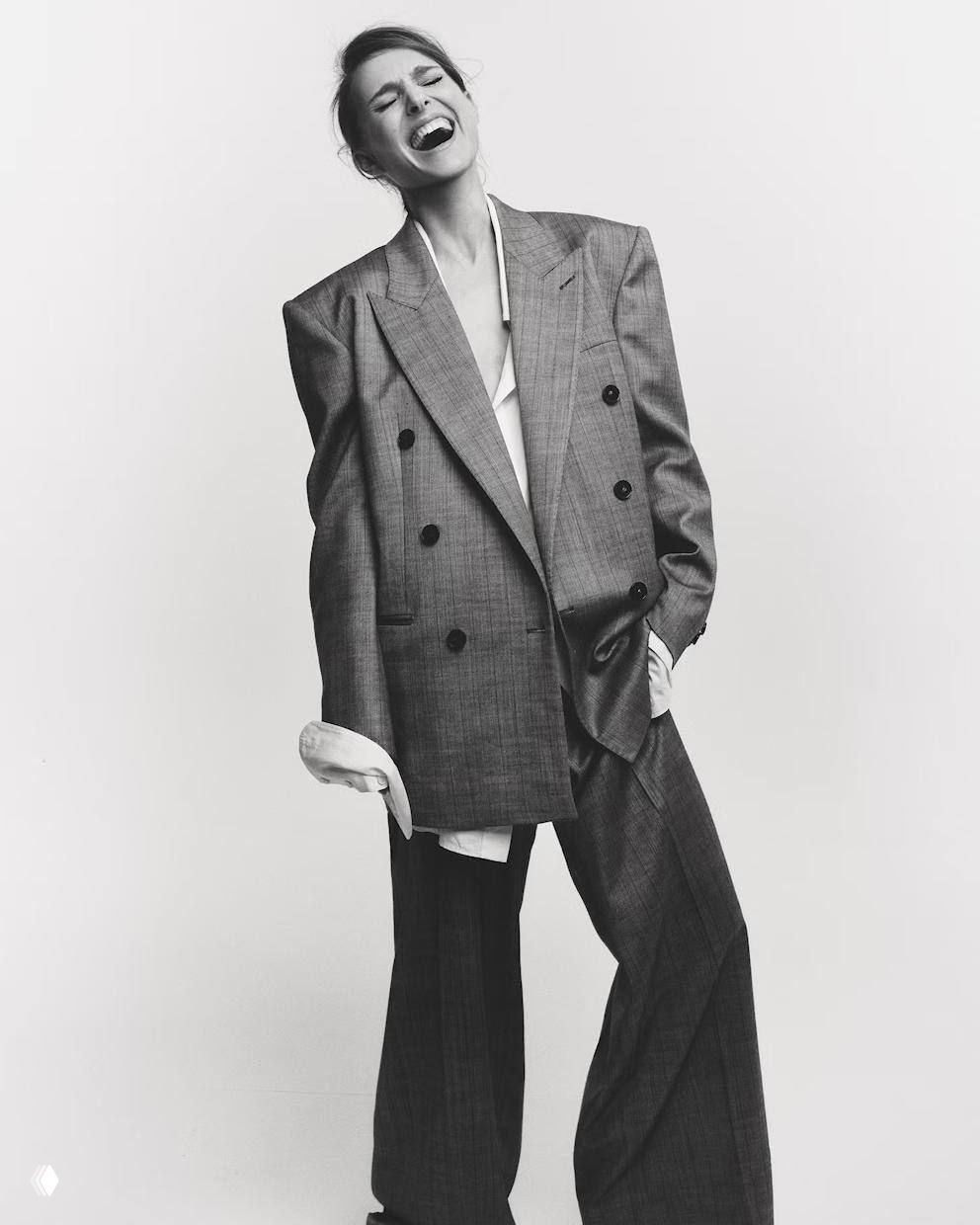Чёрно-белая студийная фотография женщины в свободном двубортном костюме и рубашке, экспрессивная поза и модный образ.