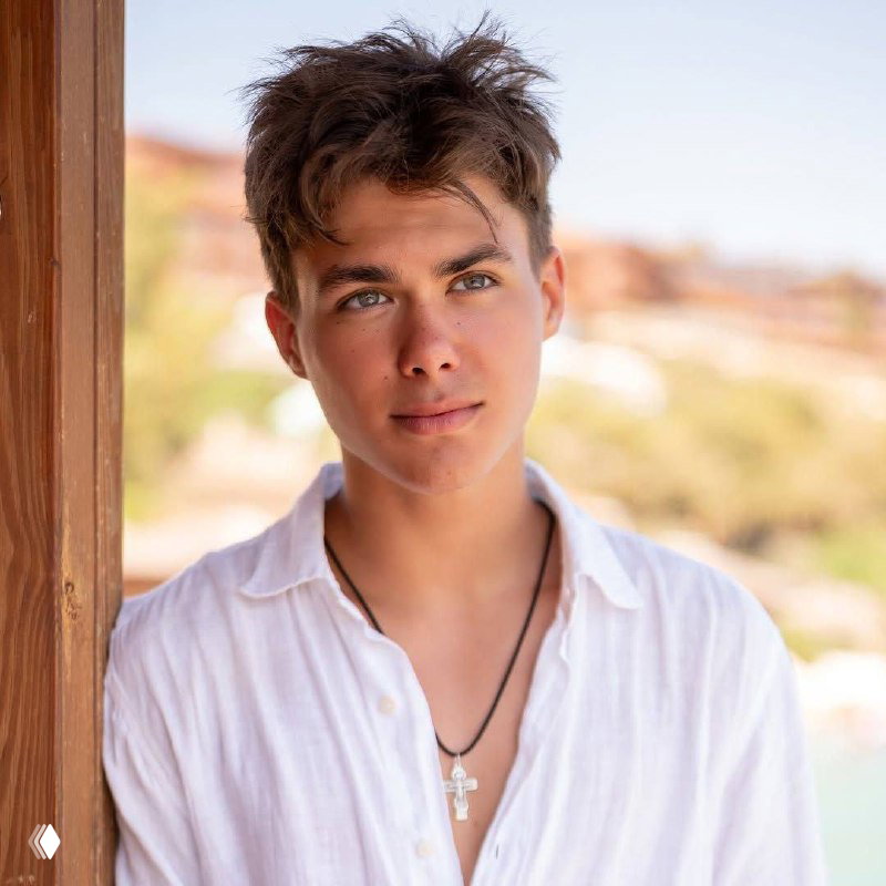 Крупный портрет подростка в белой рубашке с кулоном-крестом: мягкое боке на фоне летнего пейзажа и тёплые природные тона.