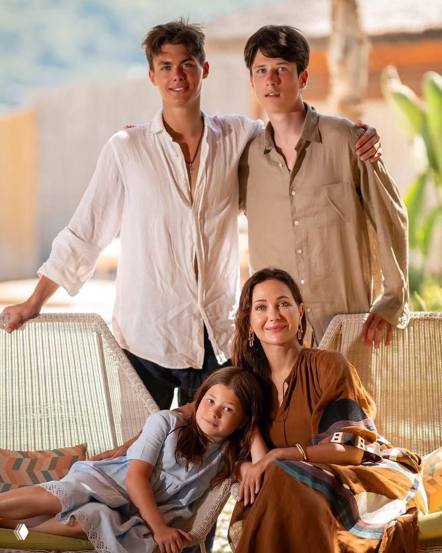 Семейная фотография на открытом воздухе: женщина сидит в кресле, рядом трое детей разного возраста в лёгкой летней одежде на террасе, тёплый свет.