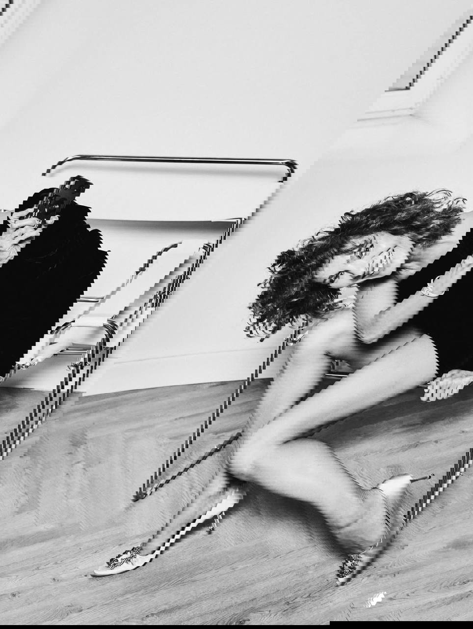 Чёрно‑белый кадр: актриса сидит в кресле, держит камеру и смеётся — динамичный, игривый момент из фотосессии.