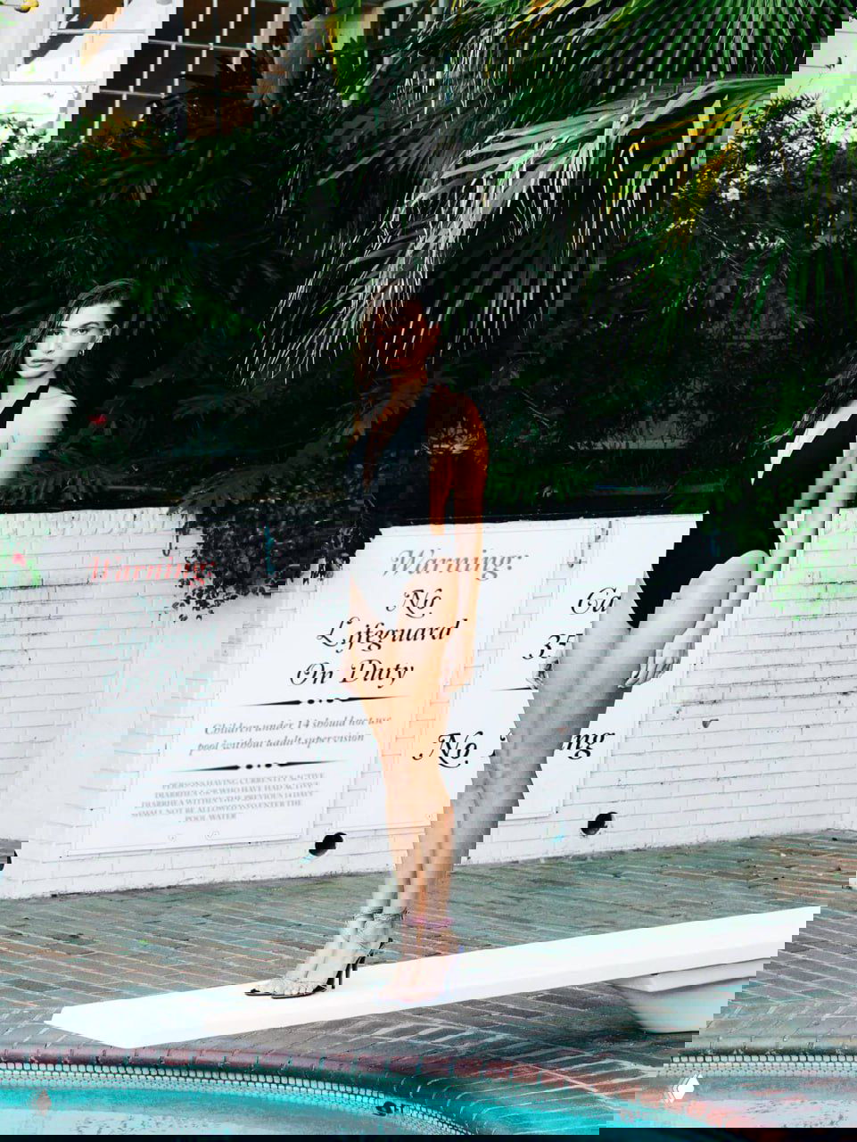 Модель стоит у бассейна в чёрном купальнике на трамплине, цветной кадр с тропической растительностью на фоне