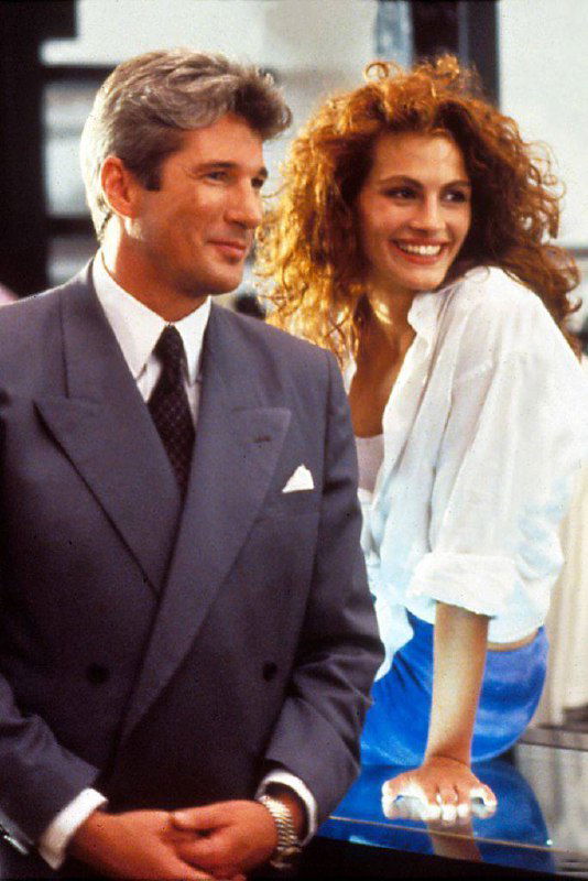 Кадр из съёмок: мужчина в деловом костюме рядом с улыбающейся женщиной с кудрявыми волосами; сцена в стиле романтической комедии/драмы.