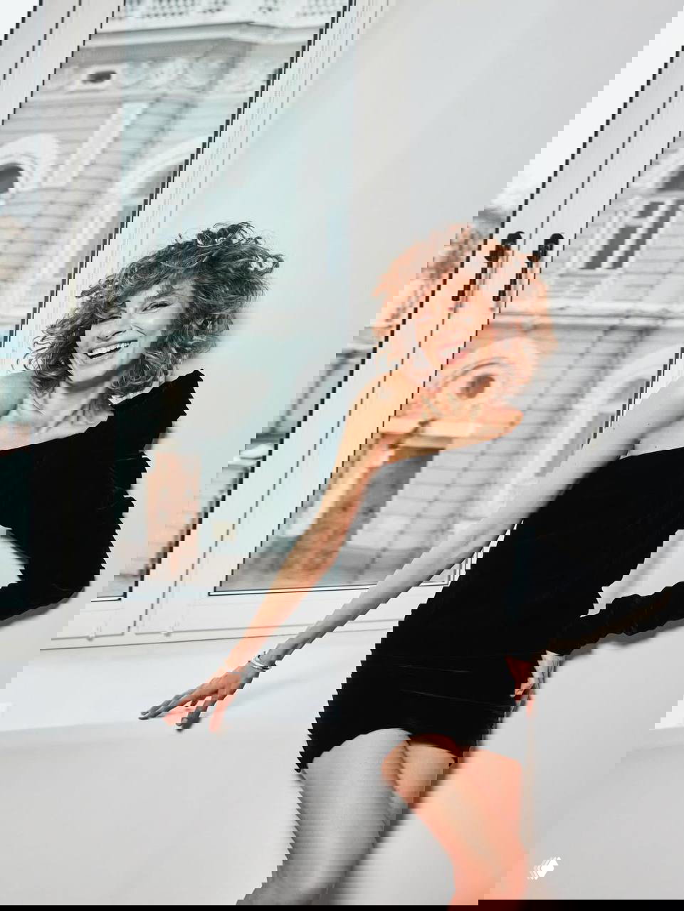 Елена Подкаминская у окна в мини‑платье — портретная фотосессия в светлом интерьере на фоне городской архитектуры за окном.