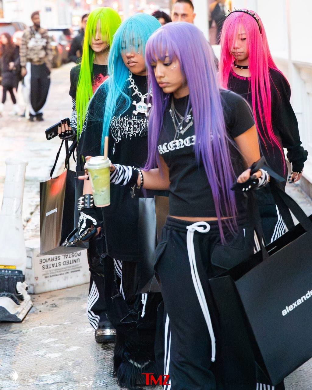 Несколько подростков в ярких париках идут по улице, одна держит напиток; на тротуаре видны прохожие и папарацци с камерами.