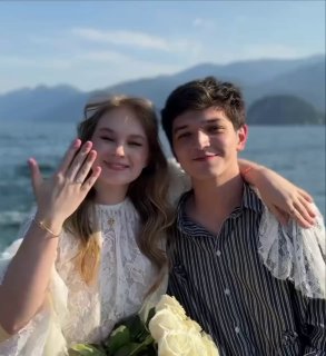 Пара на фоне моря: невеста показывает обручальное кольцо на пальце, оба улыбаются на берегу, видны вода и горы на заднем плане.