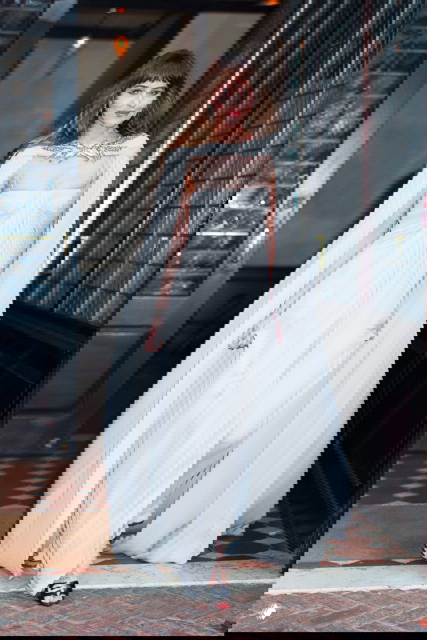 Дакота Джонсон в белом платье и длинной накидке идёт по коридору перед событием TIME100, видно полное платье и туфли на каблуке.