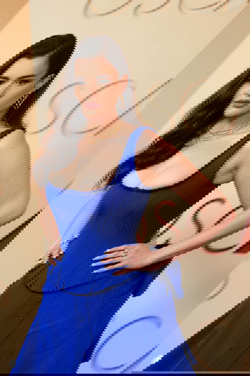 Барби Феррейра на бэкдропе церемонии Оскар 2026: руки на талии, украшения и макияж, видна надпись OSCARS и текстура синего платья.