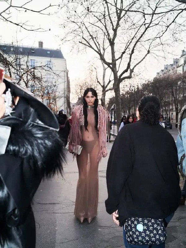 Оксана Самойлова идёт по тротуару Парижа в длинном прозрачном платье Rick Owens, вокруг толпа, городские здания и голые деревья на фоне.