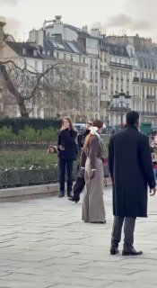 Далёкий кадр на набережной Парижа: женщина в пальто и аксессуарах идёт по тротуару на фоне классической парижской застройки и прохожих.