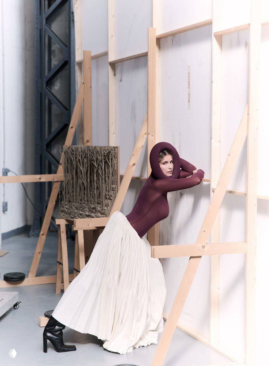 Студийный кадр: модель в бордовом облегающем верхе и длинной белой плиссированной юбке опирается на подпорки съёмочного пространства.