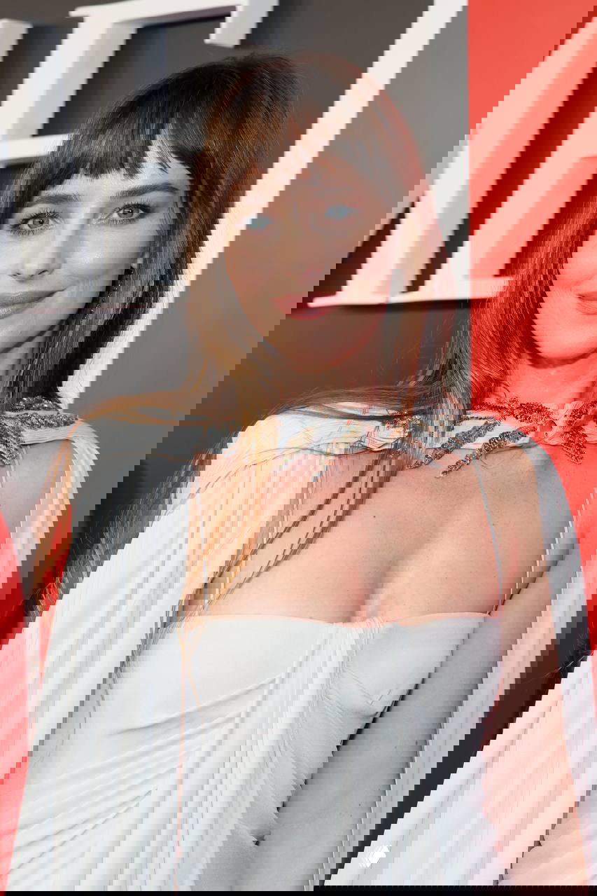 Портрет Дакоты Джонсон в белом платье с украшением у шеи, макияж и причёска видны крупным планом на фоне декора мероприятия.