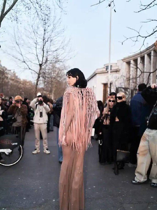 Съёмочный кадр сзади: Оксана Самойлова в прозрачном платье и бахроме, фотографы и прохожие на парижской улице, видна архитектура вокруг.