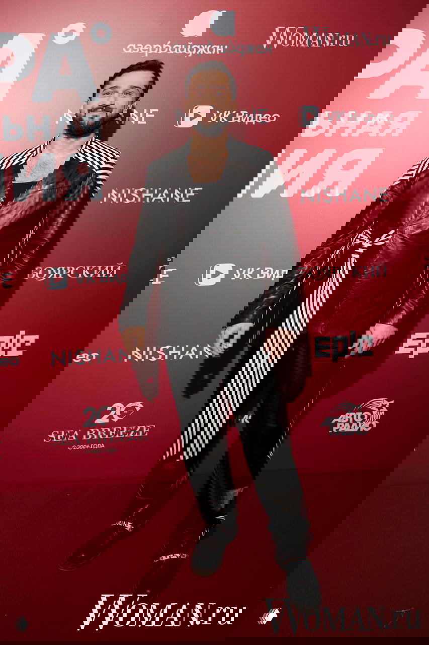 Мужчина в чёрной кожаной куртке с белыми полосами стоит на красной дорожке, фотографируется на фоне пресс-волла с логотипами спонсоров.