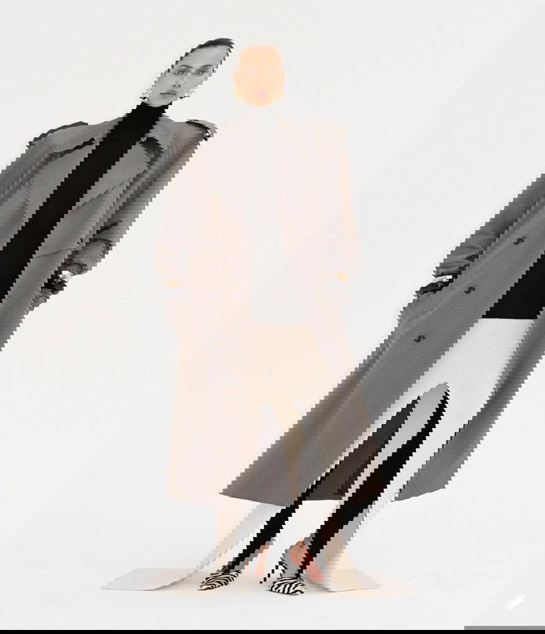 Полнофигурный кадр модели в сером тренче и белой юбке на светлом фоне, элегантный осенний образ в студийной fashion‑съёмке.