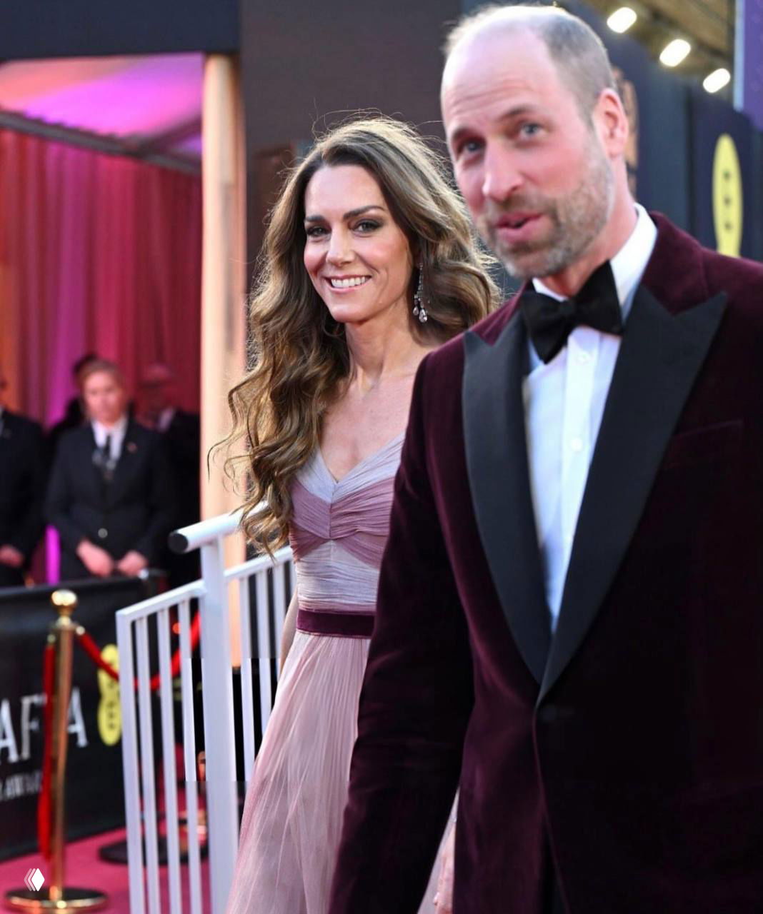 Женщина в розовом длинном платье рядом с мужчиной в смокинге на красной дорожке; кадр показывает их совместный выход на премии.