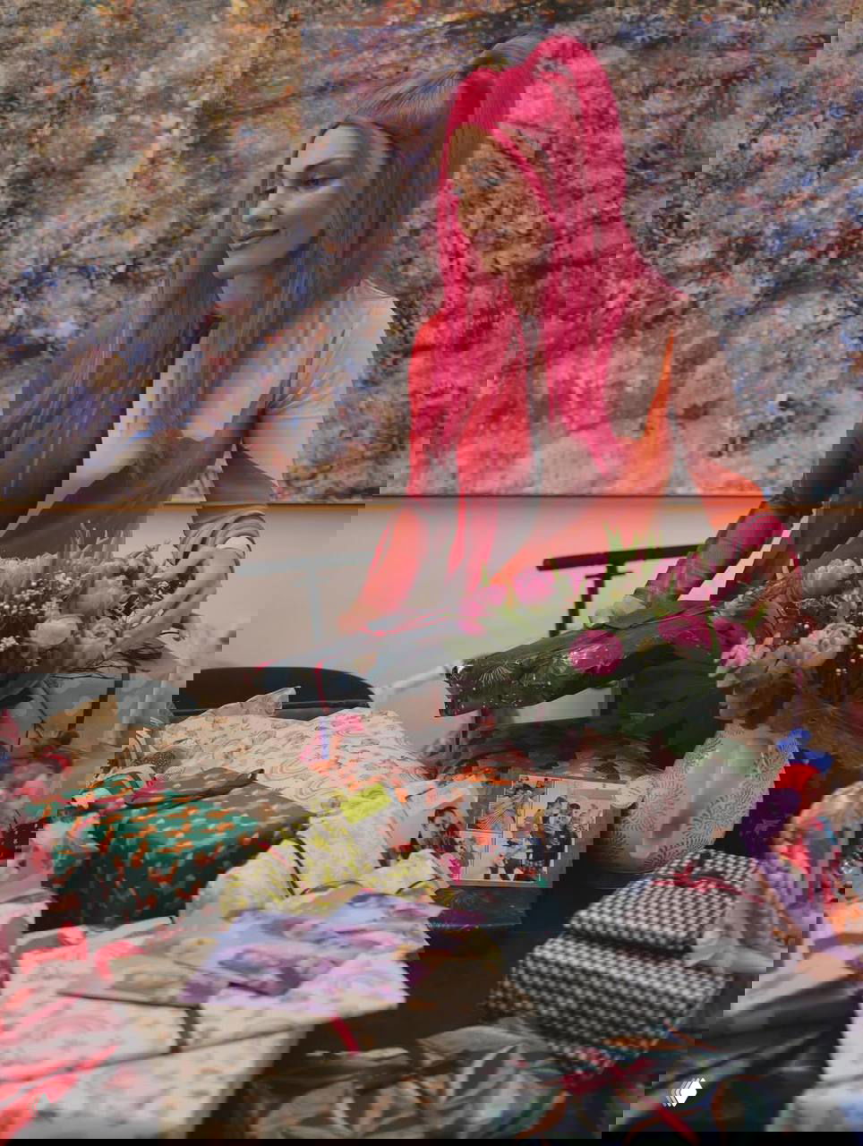 Женщина с ярко-розовыми волосами сидит за столом, рядом много упакованных подарков и букет тюльпанов — кадр с праздничного стола.