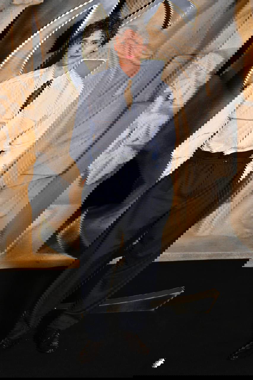 Гость в светлой рубашке и тёмных брюках с заправленной поясной линией позирует на фоне художественной инсталляции и крупного логотипа бренда YSL.