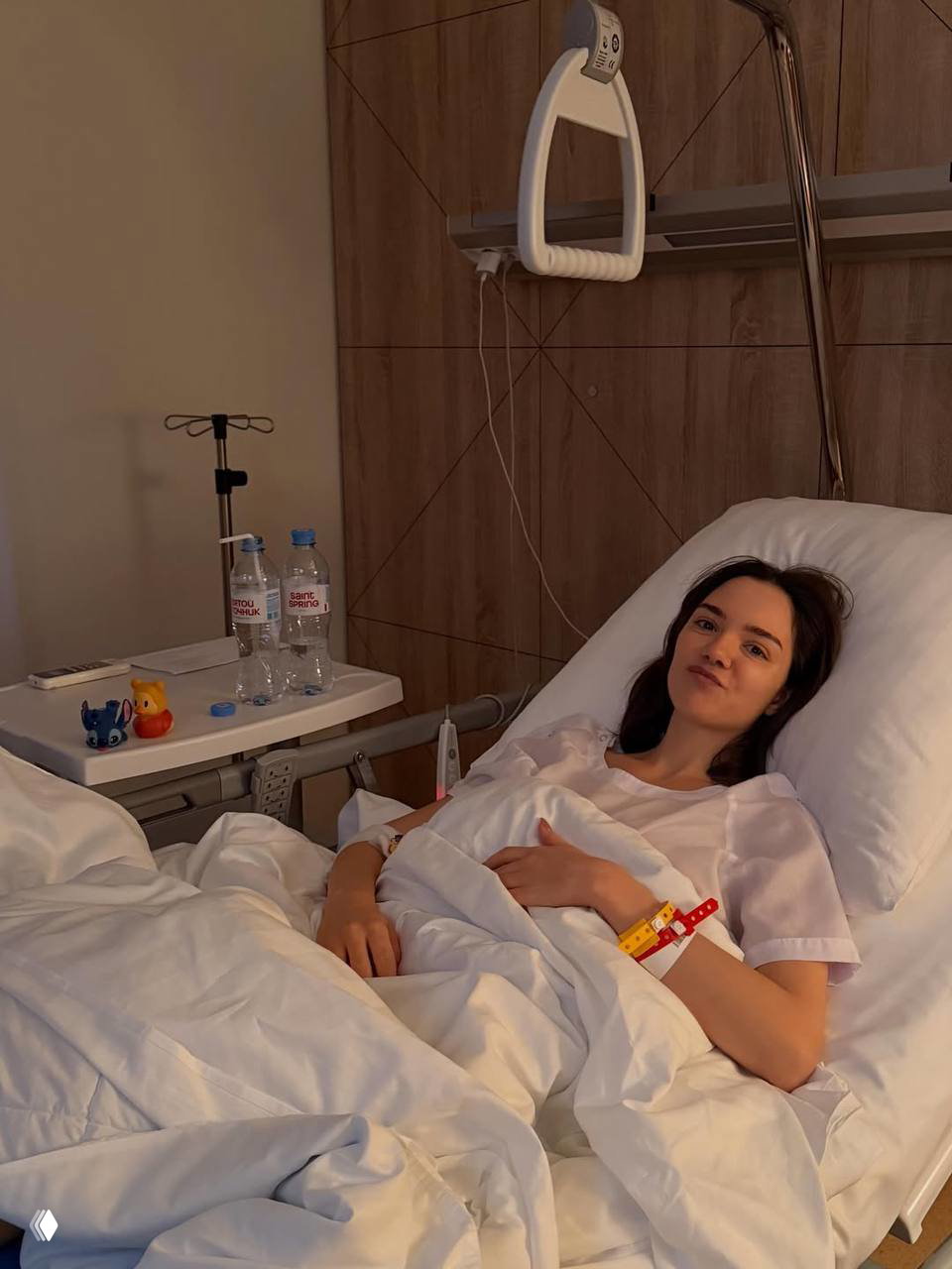 Евгения Медведева лежит в больничной кровати после плановой операции; видны постель, браслет и тумбочка с бутылками воды.