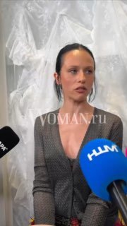 Певица Катя IOWA даёт интервью: на фоне белые ткани (возможно платья), перед лицом несколько микрофонов телевизионных каналов, серьёзное выражение лица.