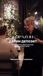 Кадр внутри зала: пожилая женщина-официантка говорит с гостями на фоне зелёного растительного декора и мягкого вечернего освещения.