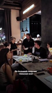 Сцена за столиками: сотрудники и посетители в ресторане во время вечера, бабушка-официантка общается с гостями, видно посуду на столах.