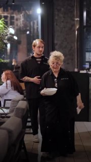 Пожилая официантка несёт блюдо через зал, рядом молодой сотрудник помогает — кадр подчёркивает харизму и рабочую атмосферу гастробистро.