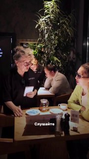 Близкий план: официантка общается с посетителями за столиком, пожилая женщина улыбается, на столе видны блюда, стаканы и салфетки.