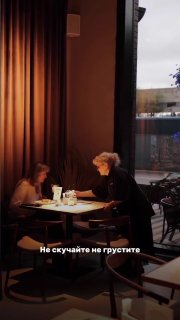 Тёплый вечерний кадр у окна: пожилая официантка разговаривает с гостями в уютной обстановке, мягкое освещение и городской пейзаж за стеклом.