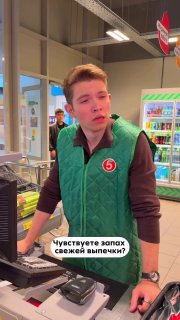 Кассир в униформе «Пятёрочка» на кассе у прилавка с товарами; на изображении видна подпись «Чувствуете запах свежей выпечки?»