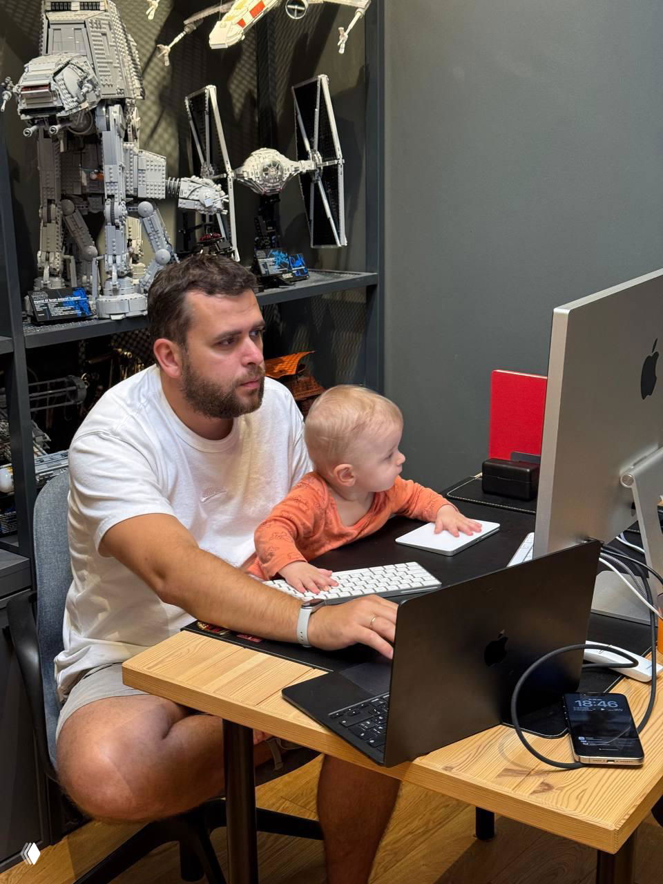 Отец и маленький сын сидят за рабочим столом у большого монитора; на заднем плане коллекция больших моделей Lego и рабочая техника — момент удалённой работы с ребёнком.