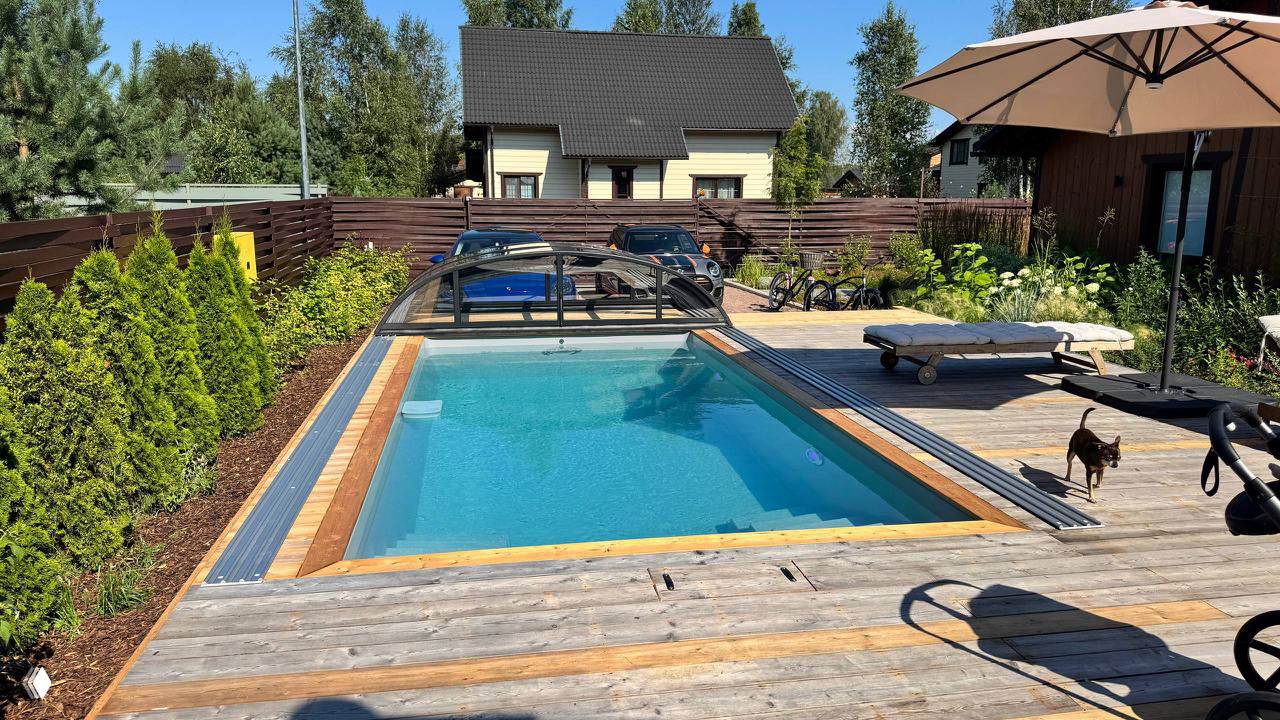 Готовый открытый бассейн на участке с деревянной террасой, навесом и котом на настиле; забор и соседние дома видны на фоне.