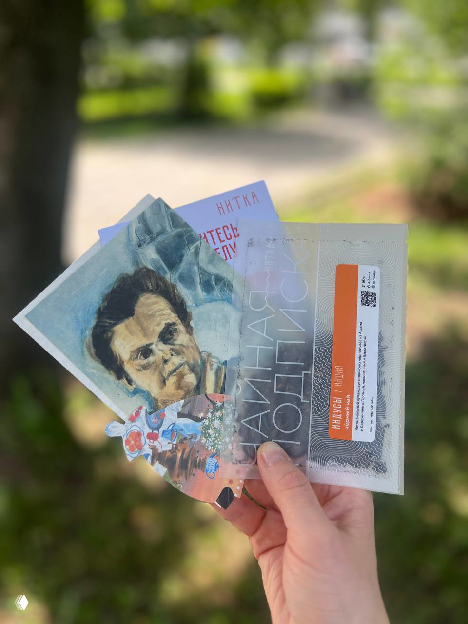 Рука держит почтовую карточку из подписки на фоне зелёного парка: размытый фон, акцент на штрихкоде и рамке открытки с дизайнерской иллюстрацией.