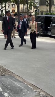 Проход гостей по улице у входа на показ Chanel: солнечный день, несколько черных автомобилей, охрана и люди с аккредитацией организаторов.