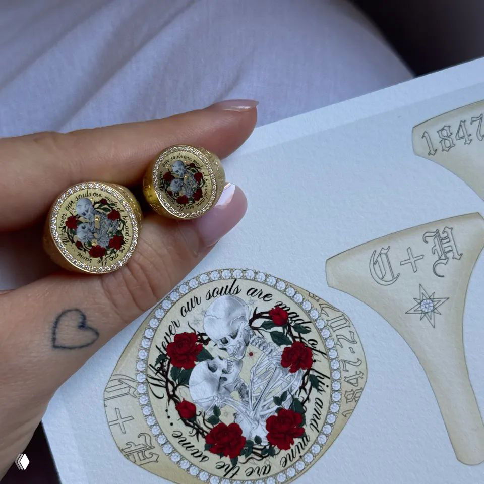 Крупный кадр парных золотых колец на бумажной зарисовке: скелеты в венке роз, видна татуировка в виде сердца на пальце.