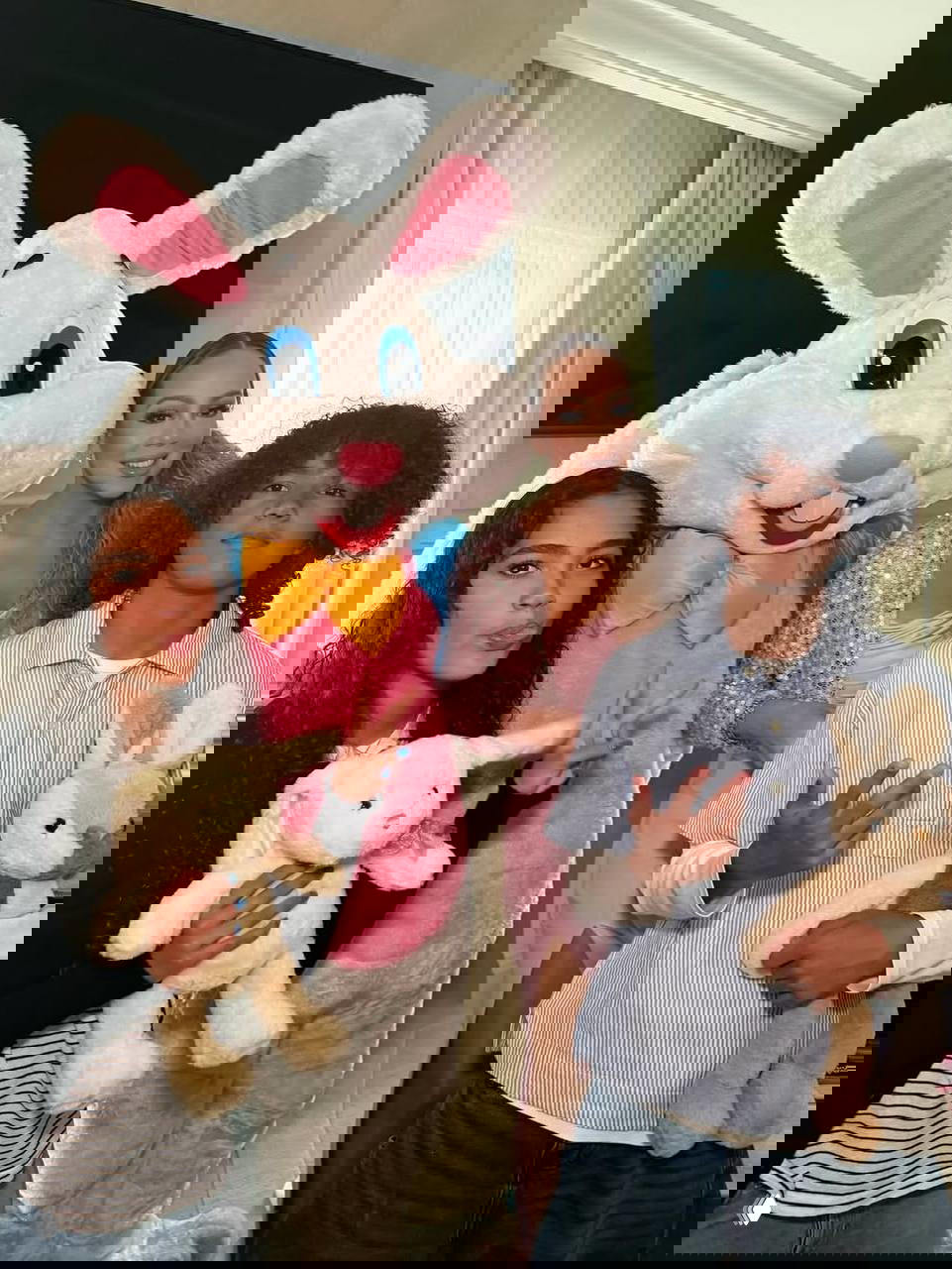 Группа людей и дети позируют рядом с ростовым костюмом зайца внутри помещения; мягкие игрушки и праздничные элементы создают атмосферу мероприятия.