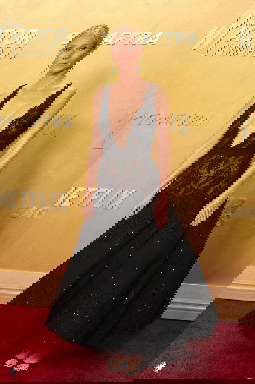 Женщина в чёрном кружевном платье с глубоким вырезом и расклешённой юбкой стоит на красной дорожке у золотой фотозоны премии.