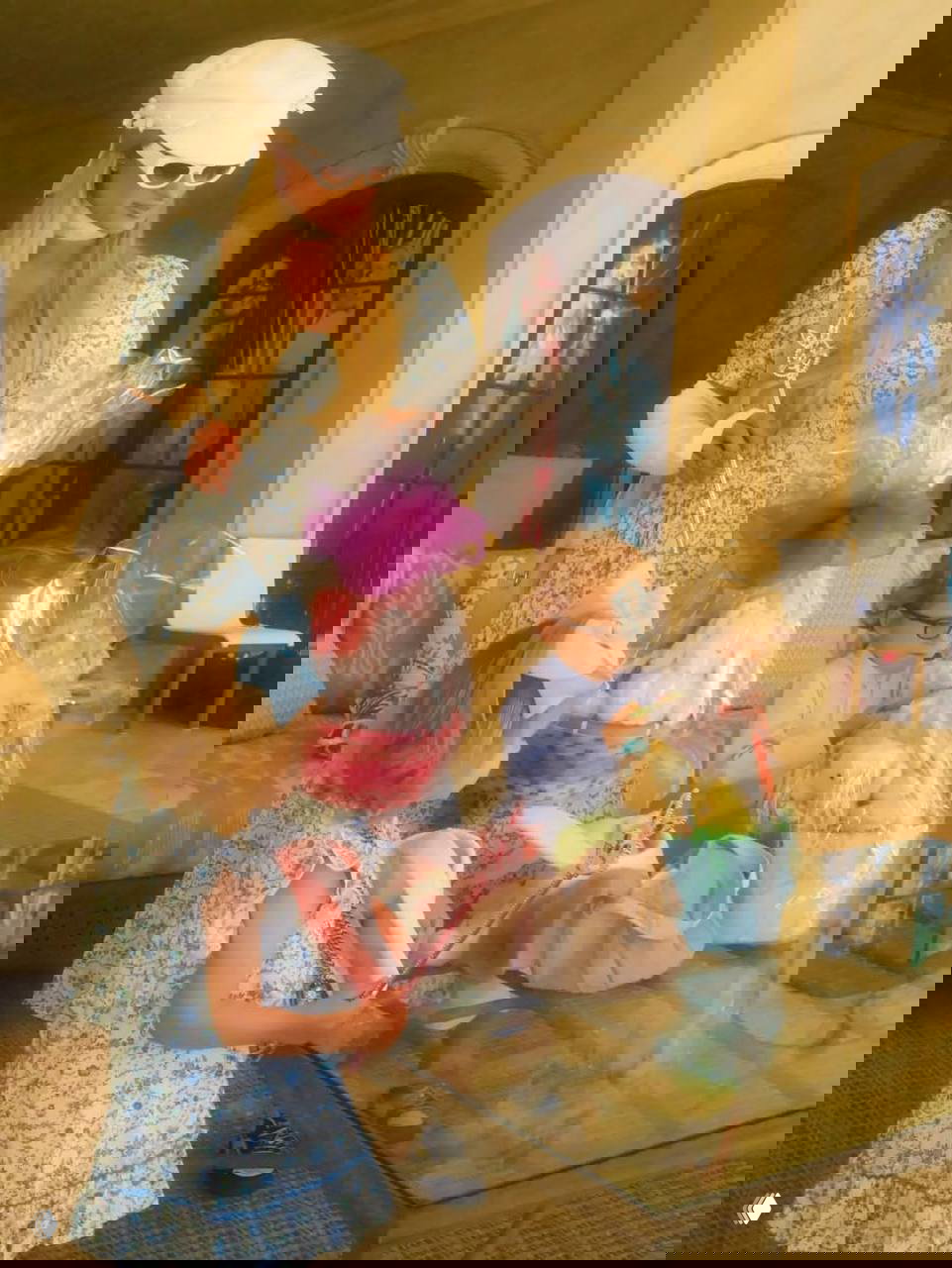 Женщина в светлом платье и дети собирают подарочные корзины и пакеты с пасхальными угощениями; интерьер украшен и готов к приёму гостей.