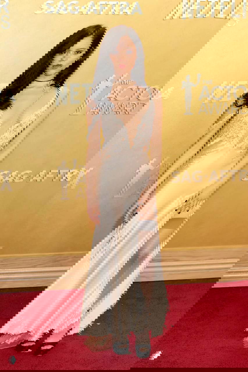 Молодая женщина в светлом атласном платье с кружевными вставками и чулком на фоне золотой бренд-стены и красного ковра SAG.