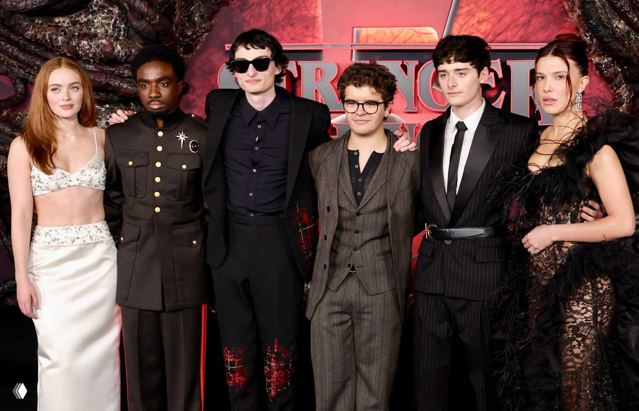 Групповое фото актеров «Очень странные дела» на премьере: несколько исполнителей ролей вместе позируют на фоне брендированного баннера Netflix