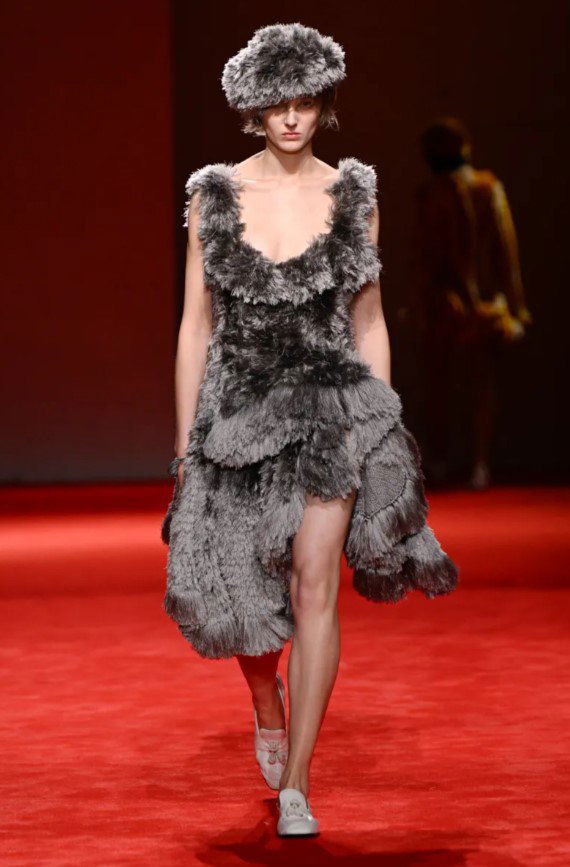 Модель в сером многослойном текстурном платье и шапке идёт по красному подиуму — фокус на тактильных фактурах и слоистом крое в коллекции.