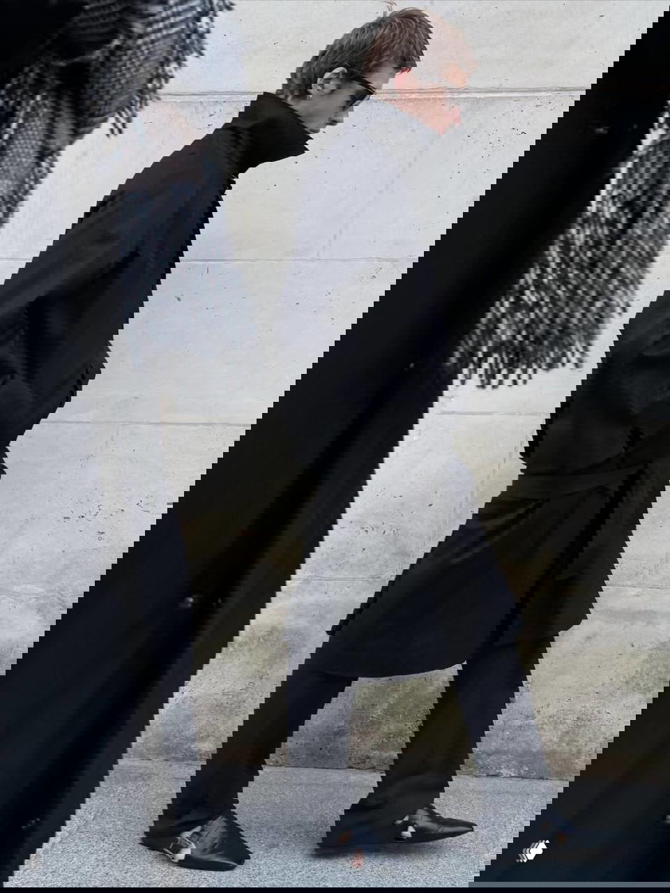 Стройная фигура мужчины в длинном пальто и солнцезащитных очках, уличная фотография в городском стиле, шаг в профиль на фоне каменной стены.