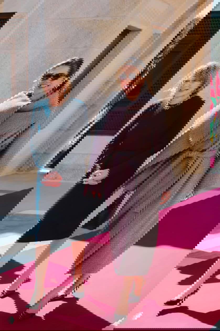 Женщина в молочно-белом брючном костюме с высоким воротником держит клатч и смотрит в сторону во время официального визита внутри здания.