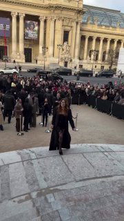 Гость в длинном чёрном пальто поднимается по ступеням перед зданием в Париже; за ней — большое скопление зрителей и фотографов.