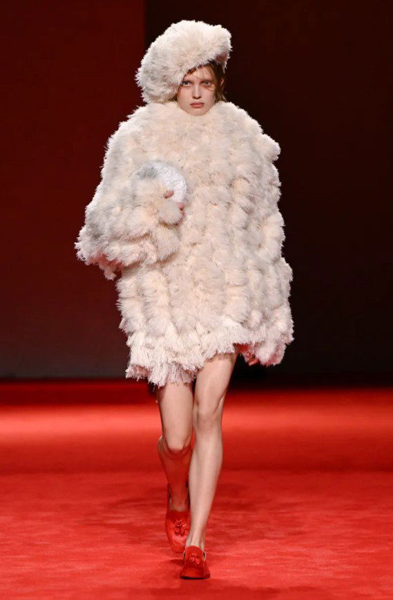 Модель в светло‑пухлой короткой шубе и шапке на красном фоне подиума, образ передаёт ощущение уюта и защитной оболочки в коллекции.